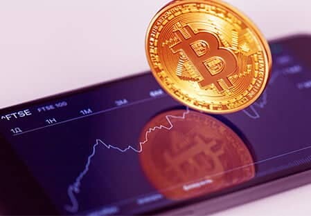 Physical gold Bitcoin coin resting over a mobile phone showing crypto asset charts